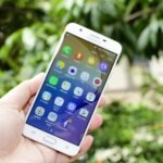 Close-up of a hand holding a smartphone displaying various app icons against a blurred outdoor background.