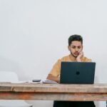 Focused young man studying on a laptop in a minimalist indoor workspace.
