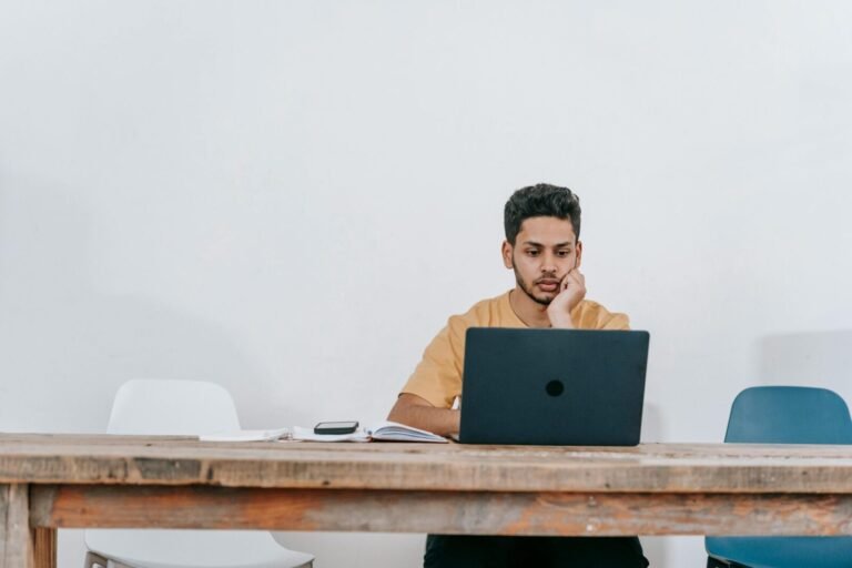 Focused young man studying on a laptop in a minimalist indoor workspace.